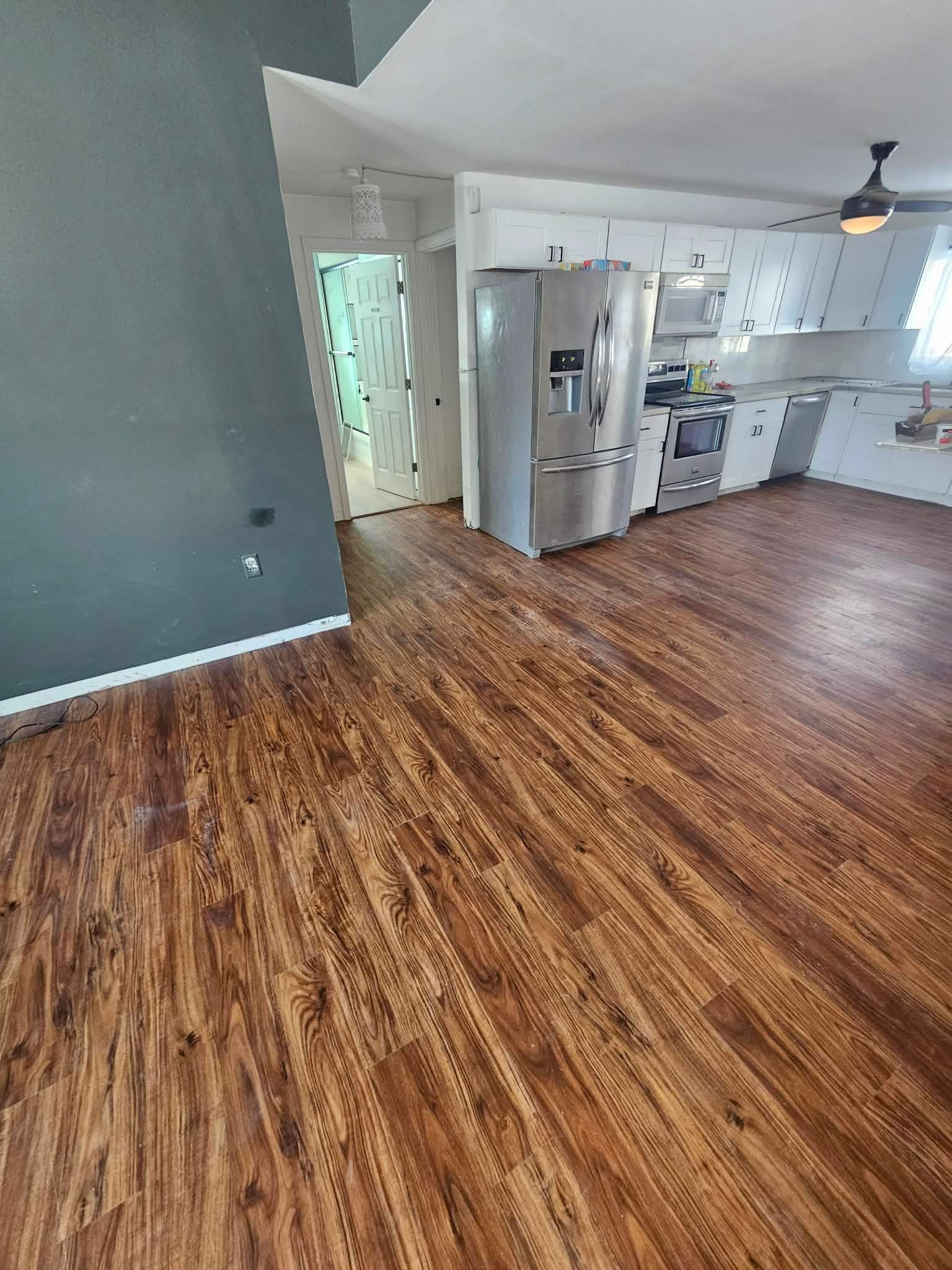 An open-plan room with wood-look flooring, a grey accent wall, and a stainless steel refrigerator in the kitchen area.