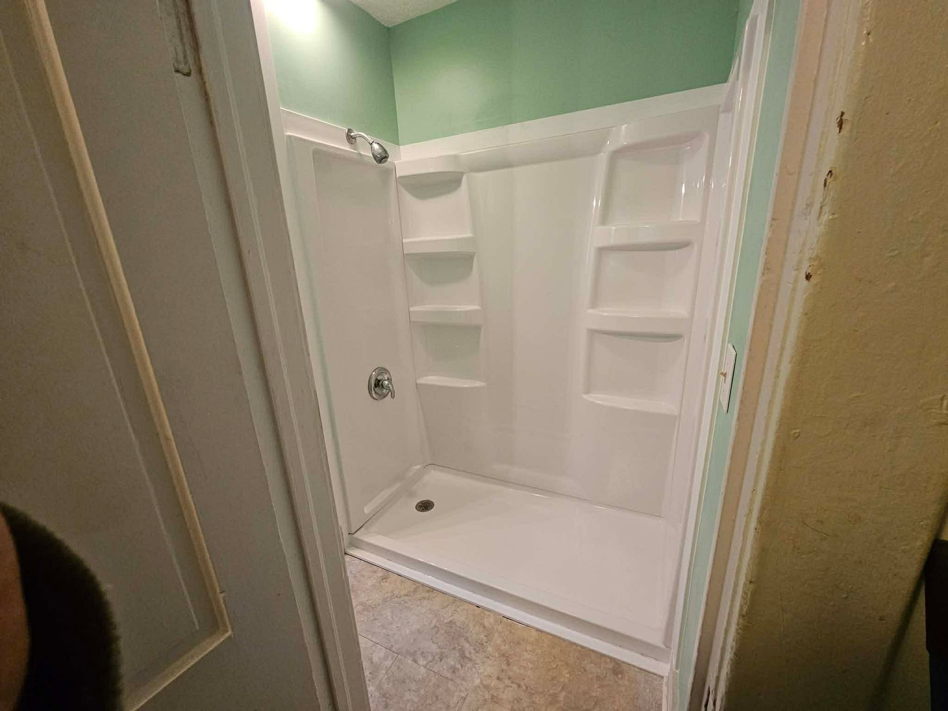 A white, three-sided shower stall with built-in shelving, set against a sage green wall in a bathroom.