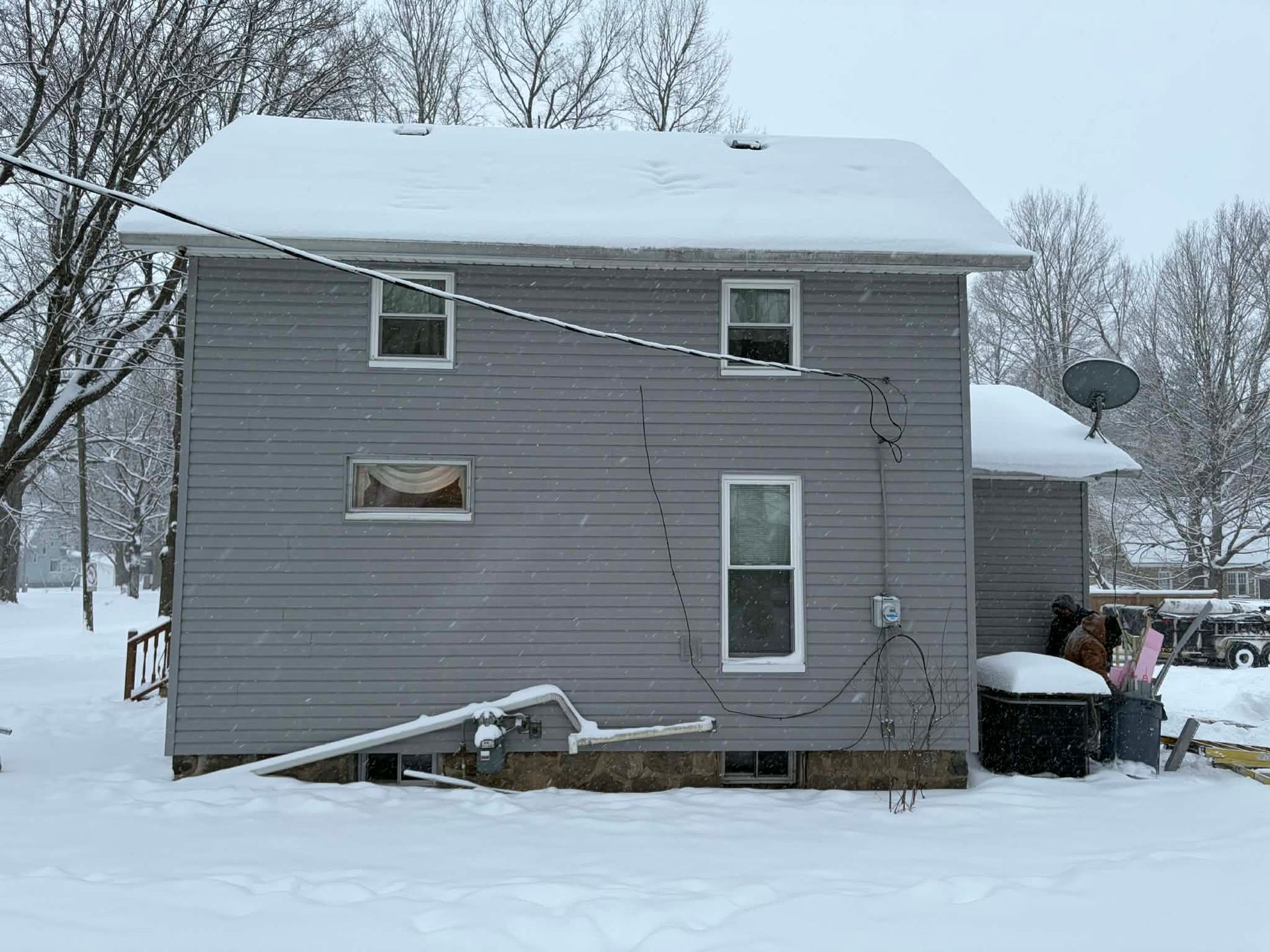 A two-story grey house exterior covered in light snow, with several windows, exposed wiring, and a satellite dish.