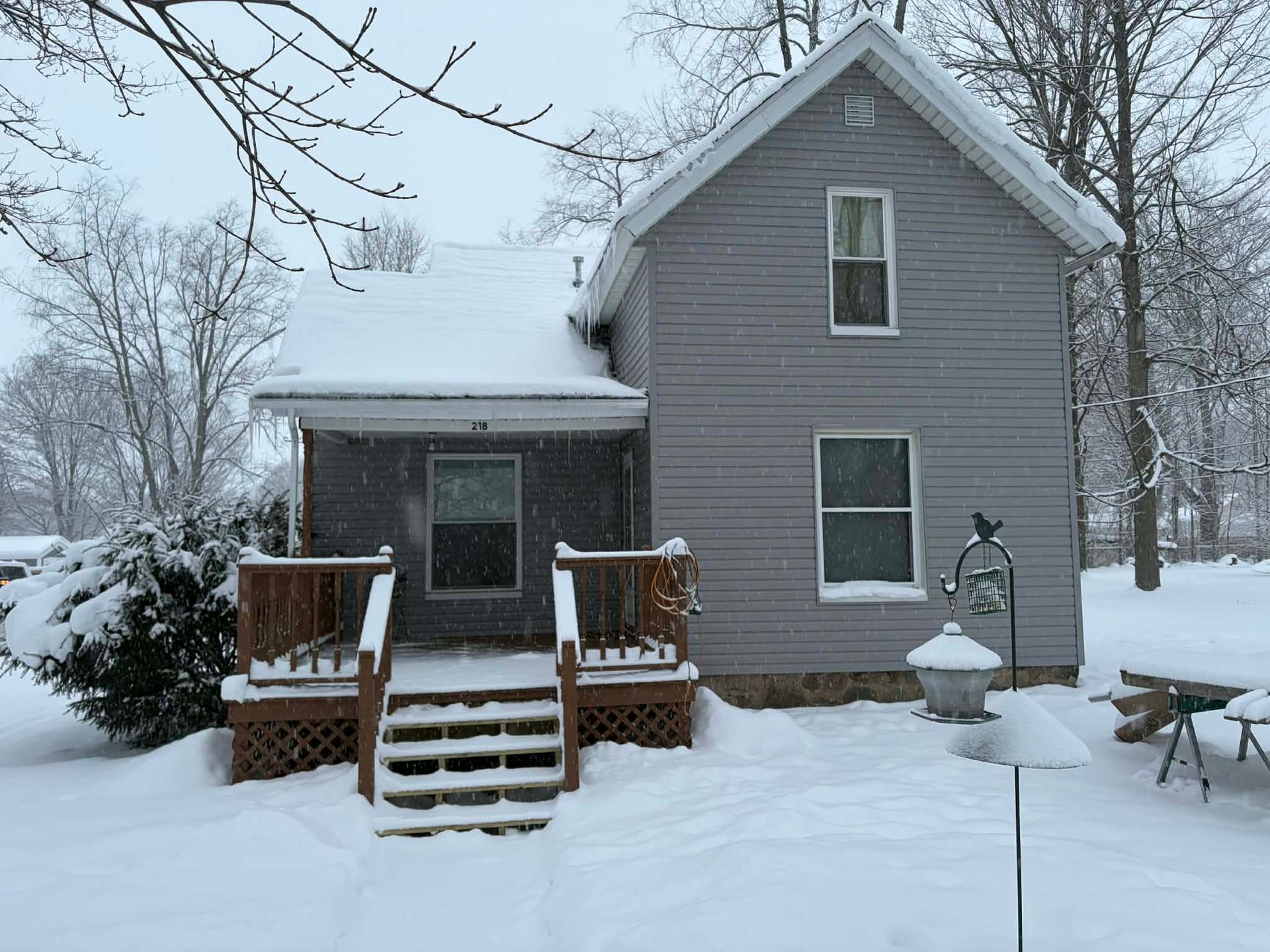 A gray two-story house with a wooden porch covered in deep snow on a winter day.