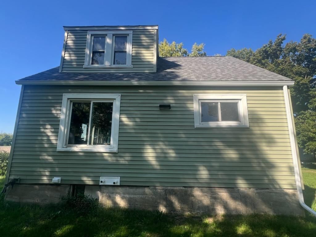 Light green house with a dormer window, white window trim, and a concrete foundation on a sunny day.