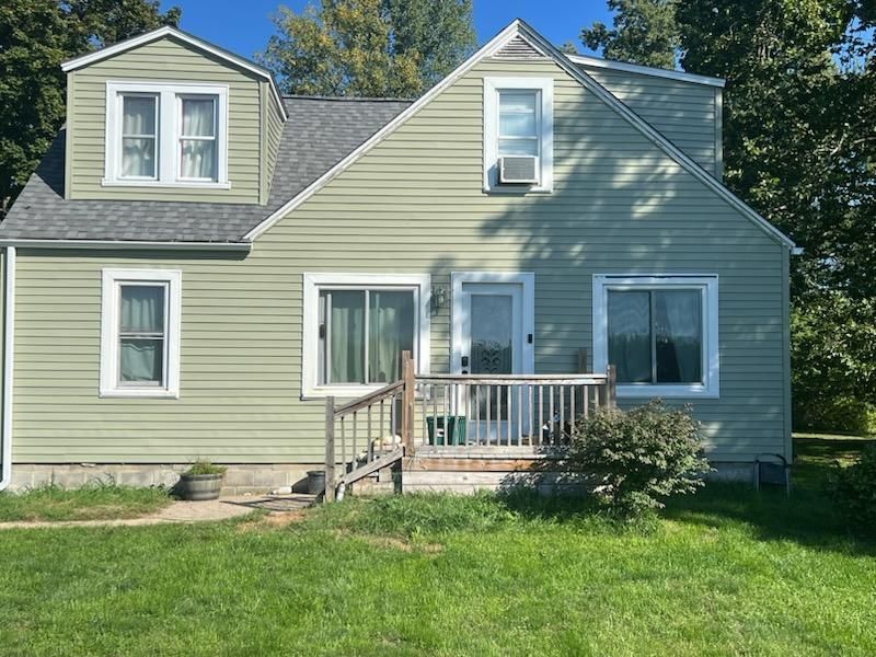 A light green, two-story house with a wooden front porch, grey shingled roof, and green lawn under a clear blue sky.