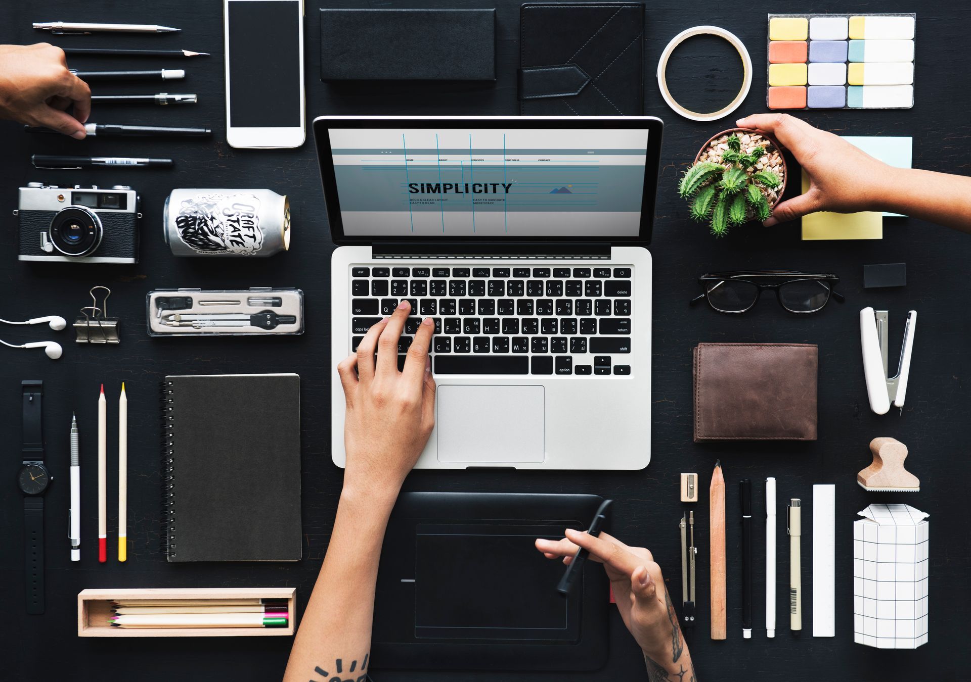 Person typing on laptop, surrounded by office supplies and a small plant, on a dark surface.