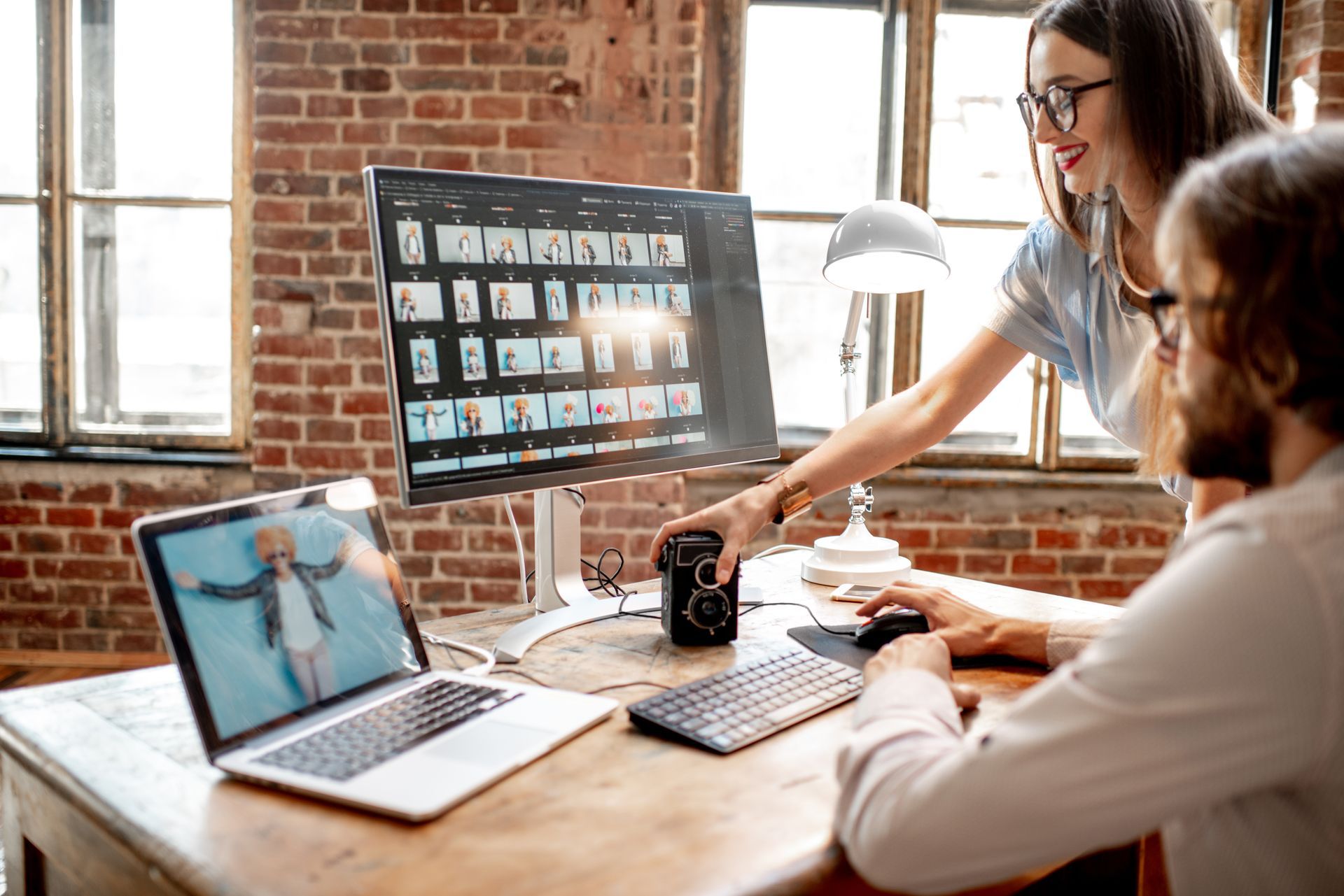 Two people reviewing photos on a computer screen and laptop in a brick-walled office.