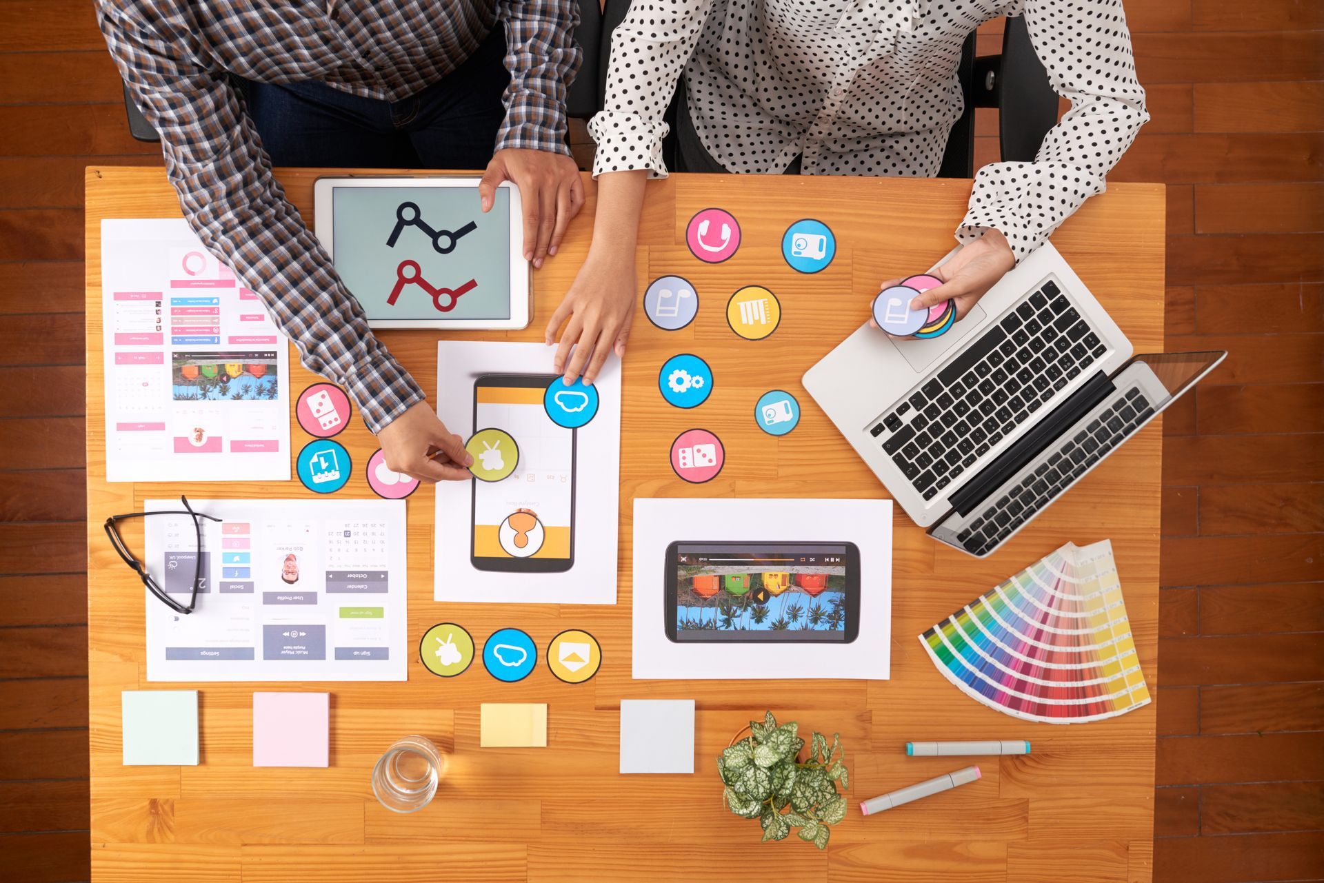 Two people collaborating at a wooden table, working on app design. The table displays a laptop, tablet, design mockups.