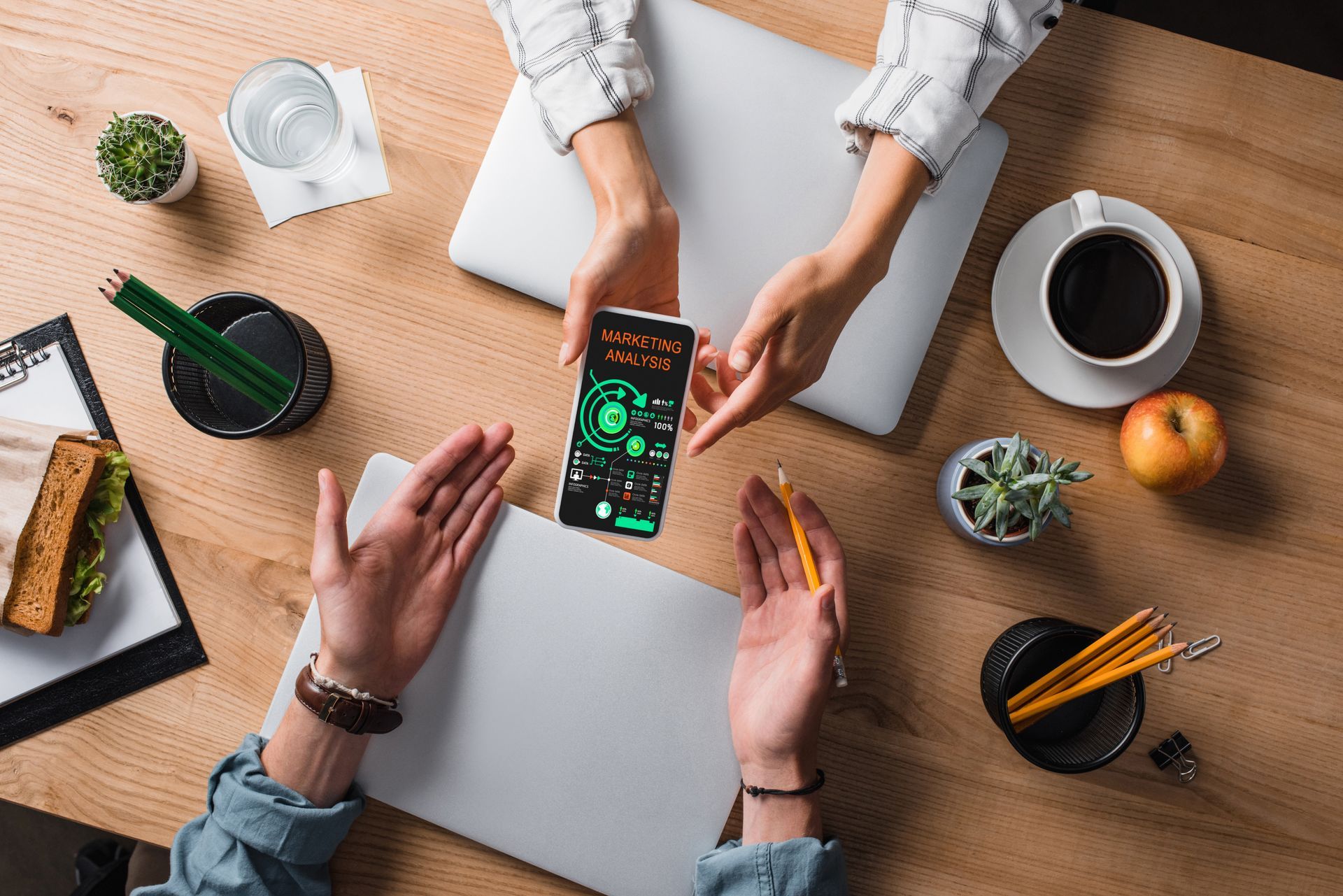 Two people share a phone with a green interface above two laptops on a wooden desk with a sandwich, coffee, and plants.