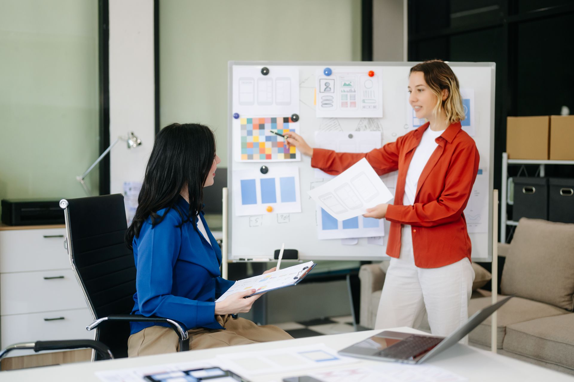 Two women discussing a presentation on a whiteboard. One points, the other holds papers. Modern office setting.