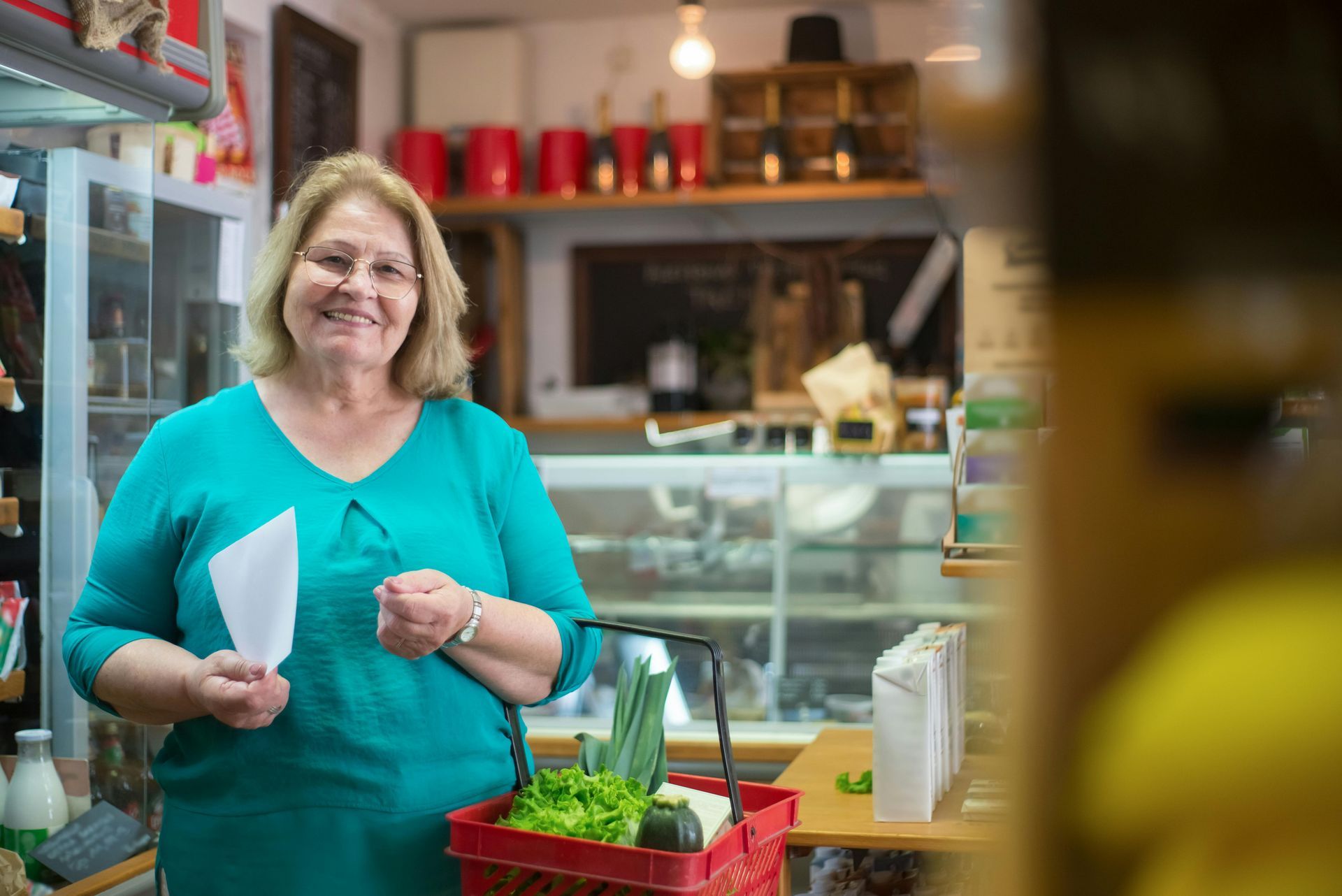 Woman in a teal shirt smiles in a shop, holding a basket of produce and a paper.