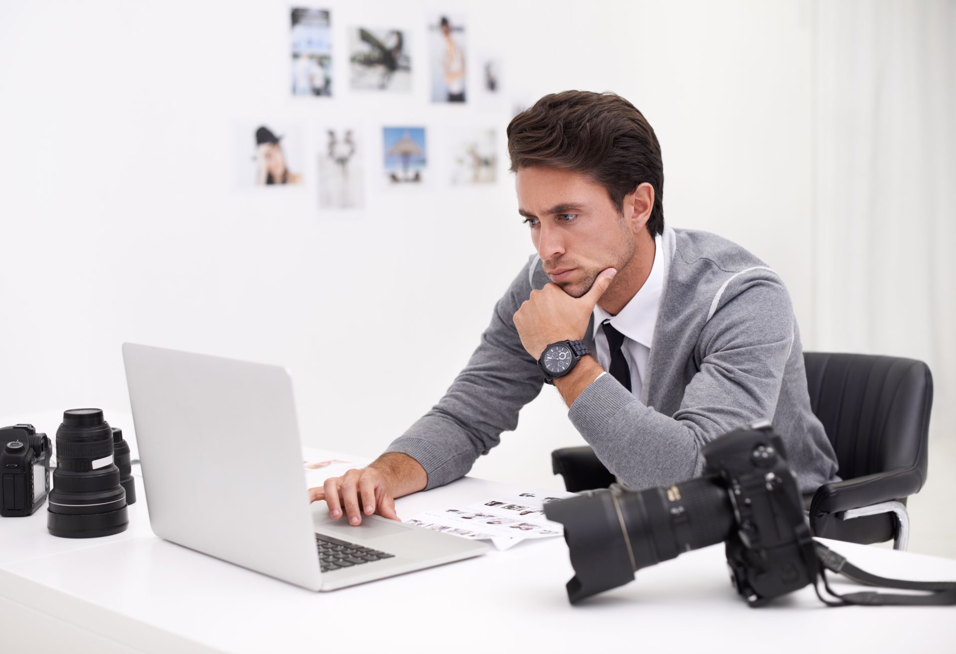 A man with a watch and sweater looks at a laptop. Cameras and photos are on the white desk.