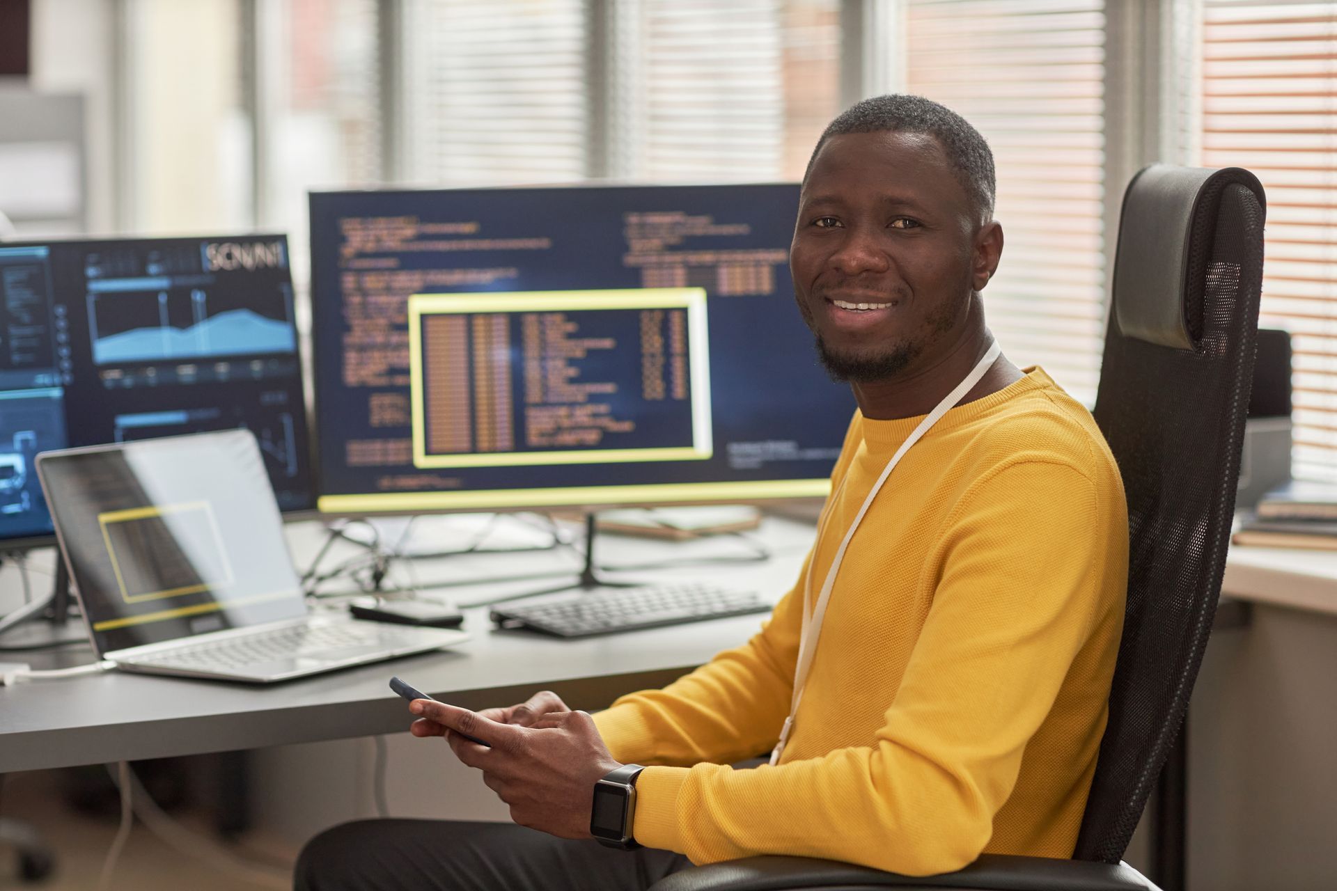 Man smiling, sitting at desk with laptop and multiple monitors displaying code in a modern office.