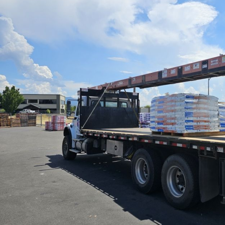 A white flatbed truck parked outdoors on an asphalt lot, loaded with pallets of materials under a bright, cloudy sky.