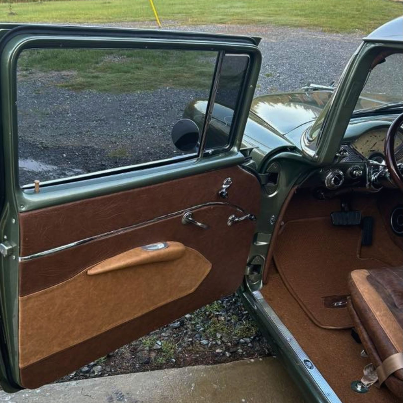 The open driver-side door of a vintage green car with a two-tone brown interior and plush fabric door panels.