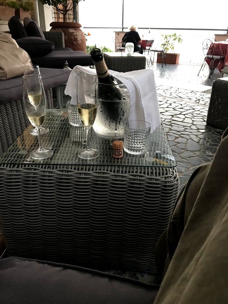 Bottiglia di champagne nel secchiello del ghiaccio, bicchieri di spumante sul tavolo all'aperto con figure in lontananza e vista sull'acqua.