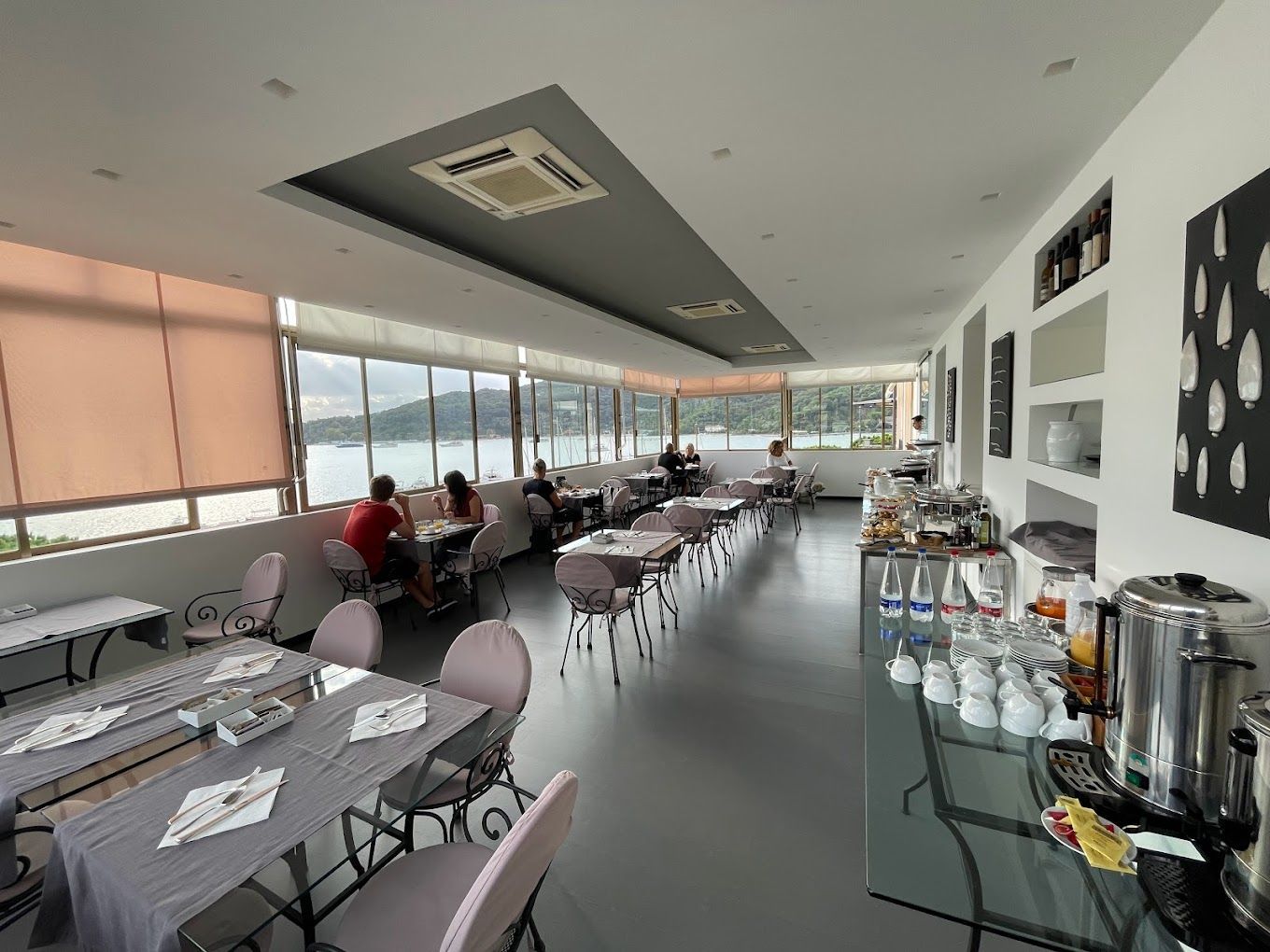 Interno di un ristorante con tavoli apparecchiati per cenare vicino a grandi finestre che si affacciano su uno specchio d'acqua.