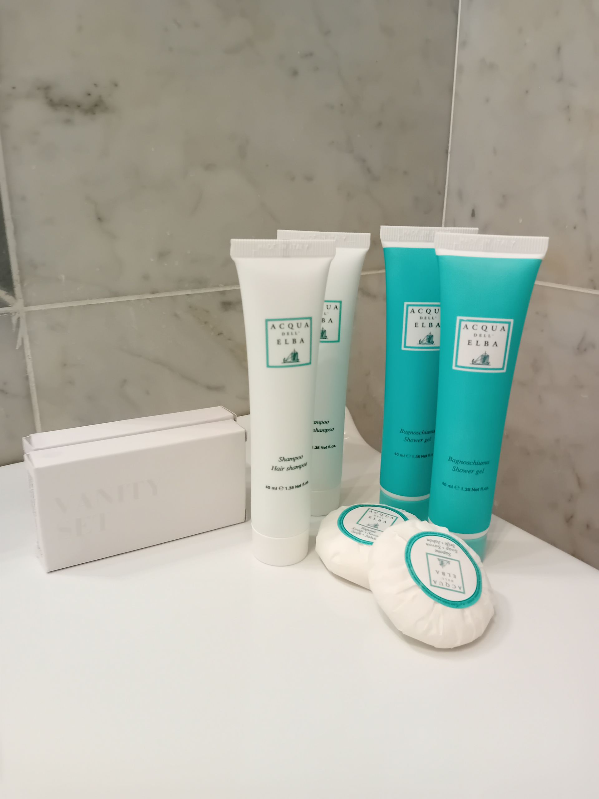 Articoli da toeletta da bagno su un bancone bianco. Tubetti azzurri e bianchi, saponi incartati e una scatola. Parete piastrellata grigia.