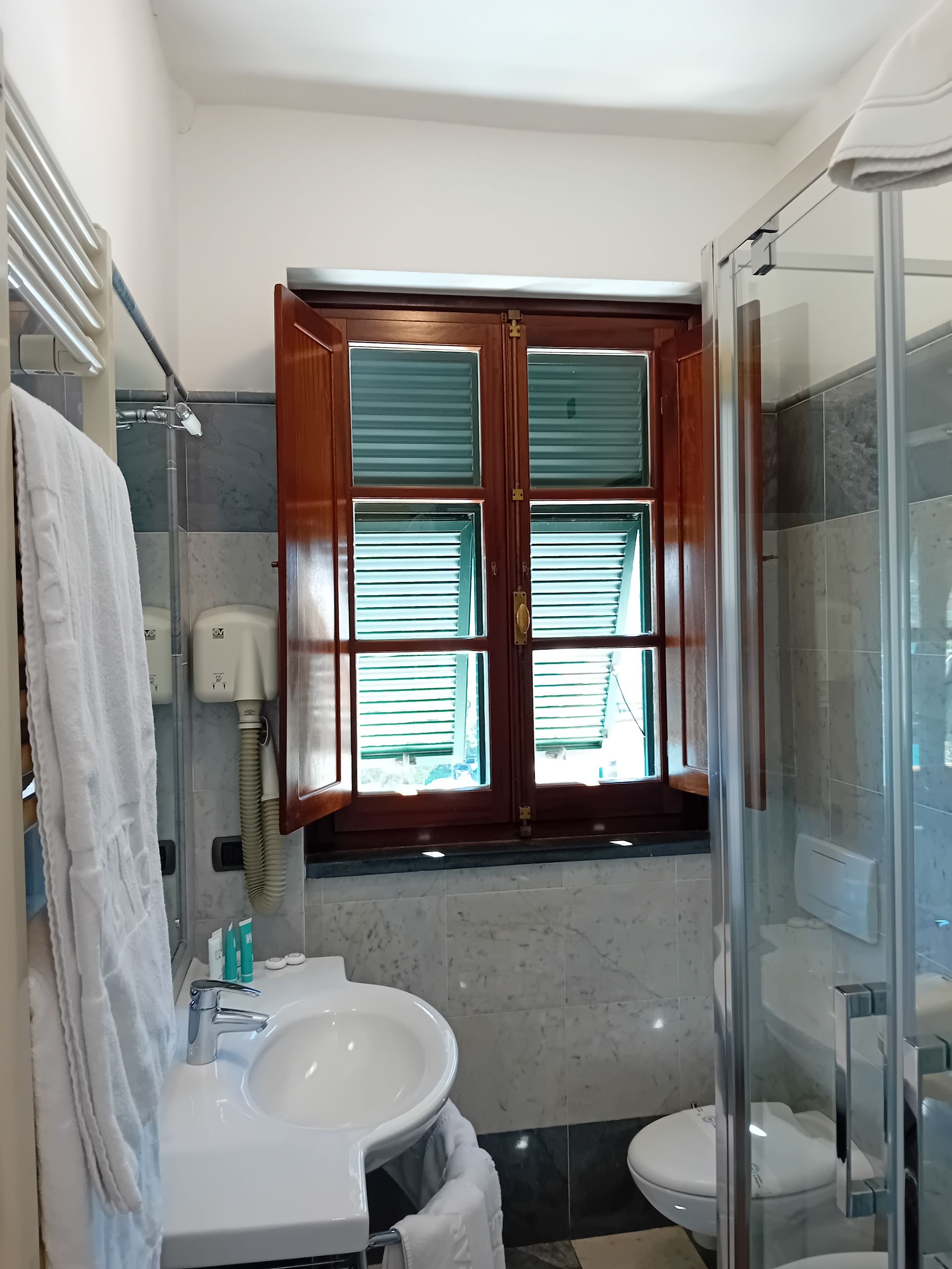 Bagno con lavandino bianco, WC, doccia e finestra con persiane aperte.