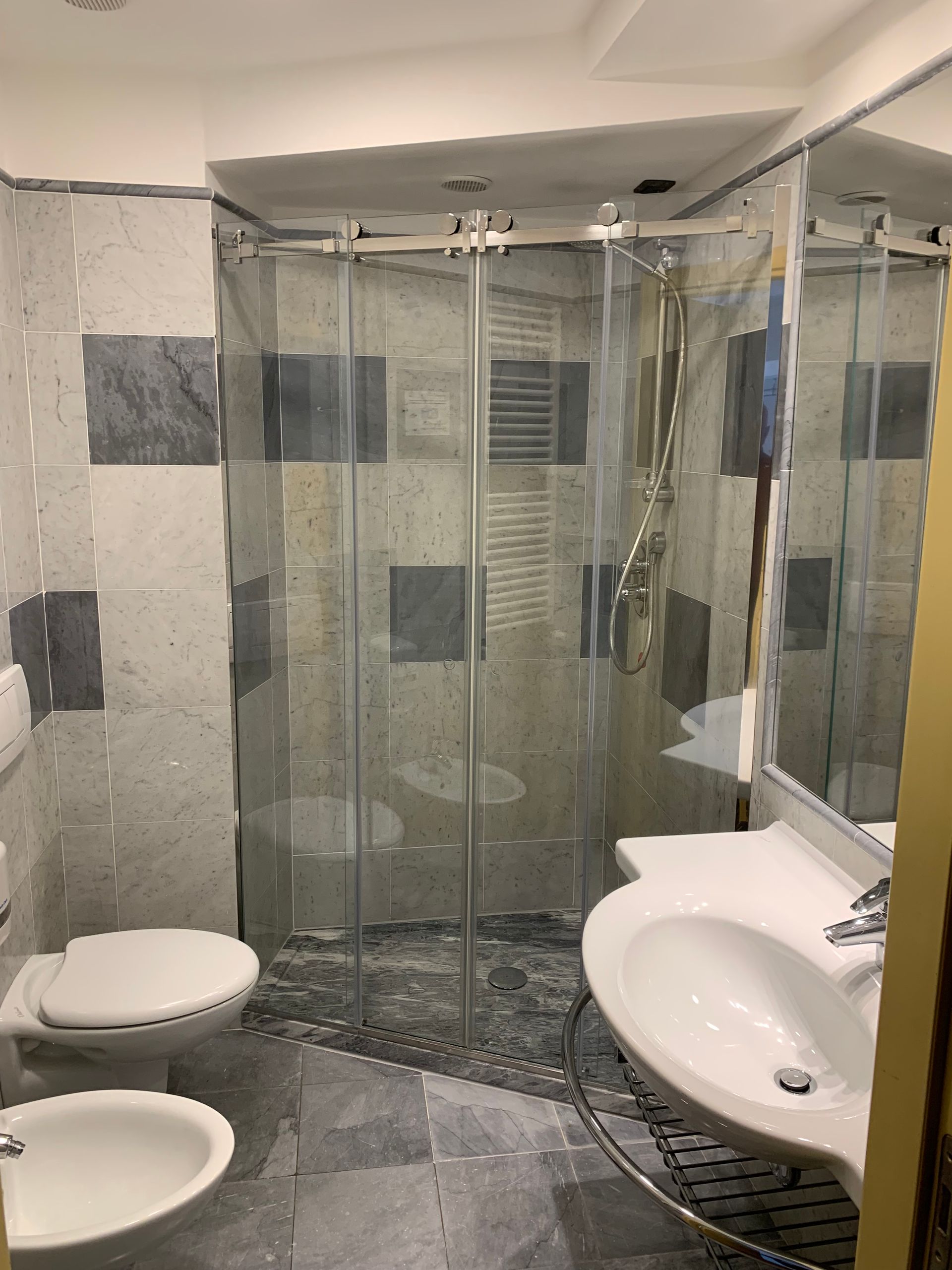 Piccolo bagno con WC, bidet, lavandino e doccia. Piastrelle grigie e bianche, porte della doccia in vetro.