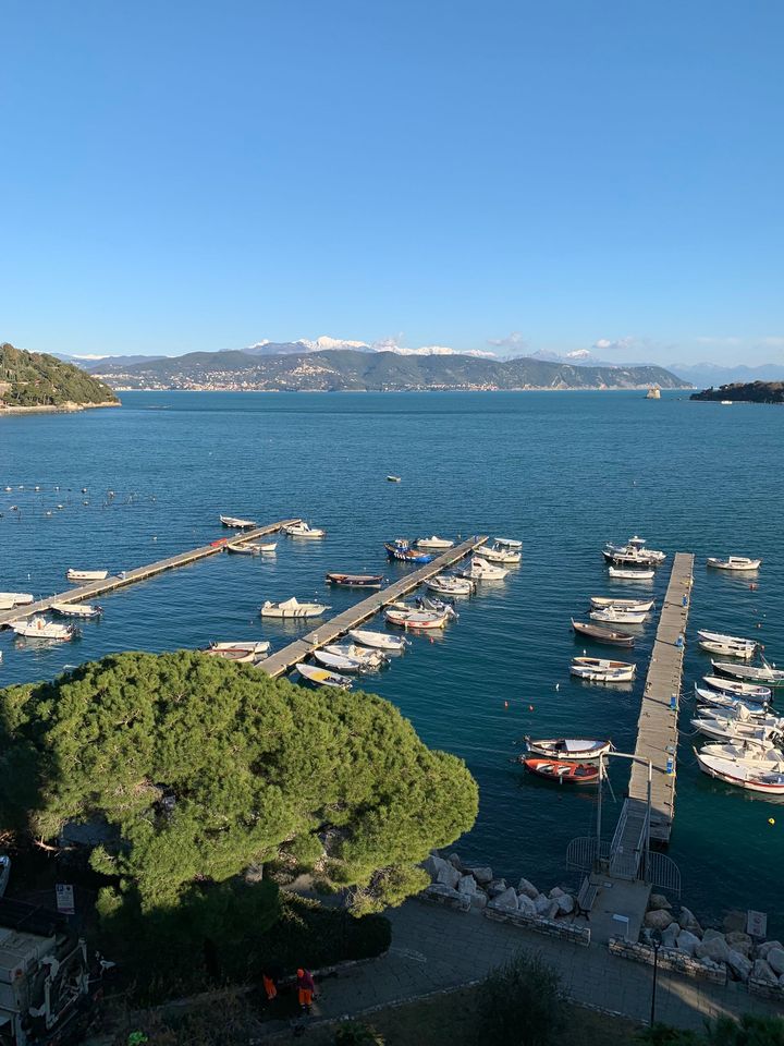 Barche attraccate ai moli in una baia azzurra, con montagne innevate in lontananza sotto un cielo limpido.