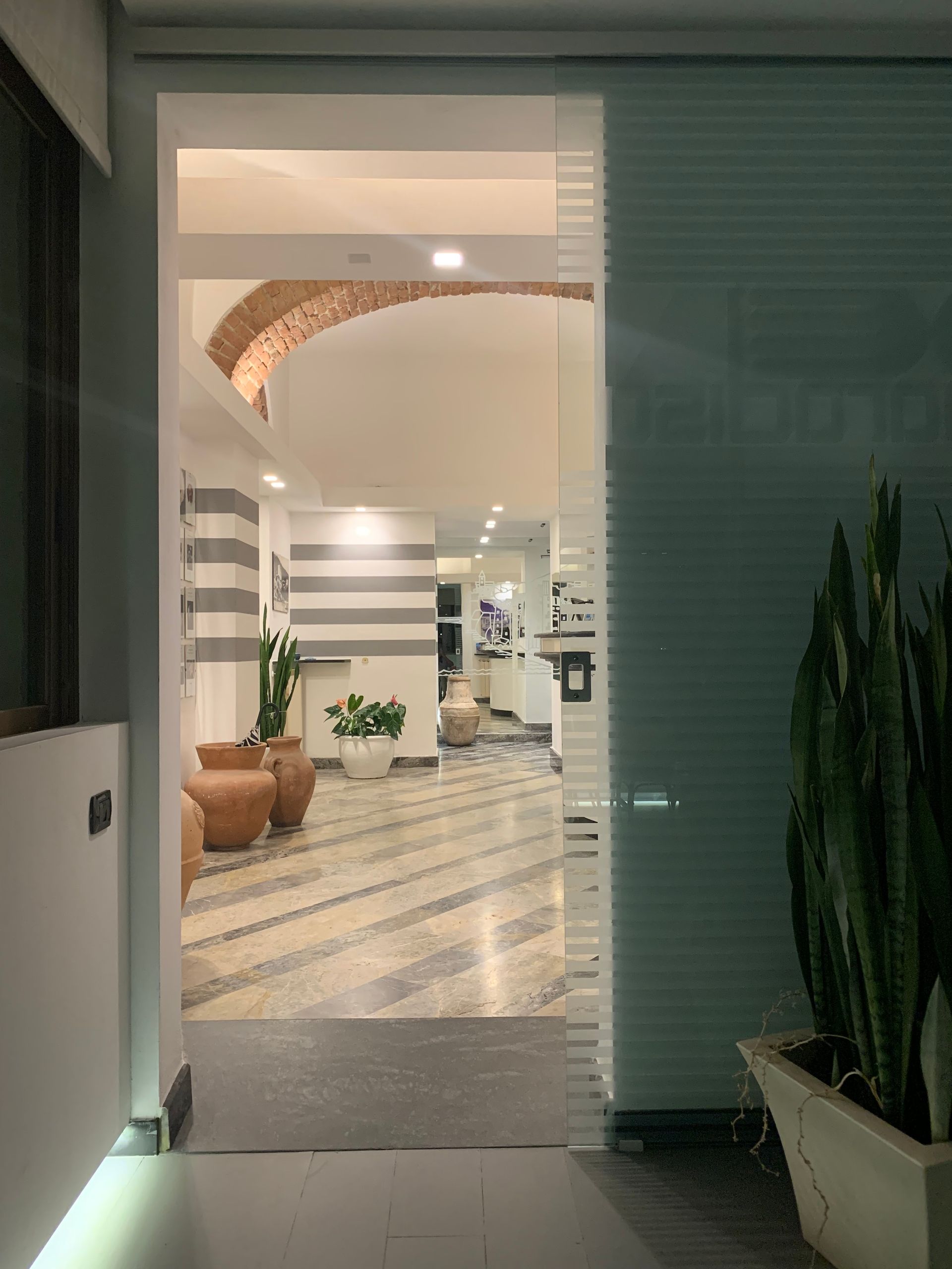 Ingresso in un corridoio con parete a strisce, pavimento a mosaico e piante. Attraverso una porta in vetro smerigliato.