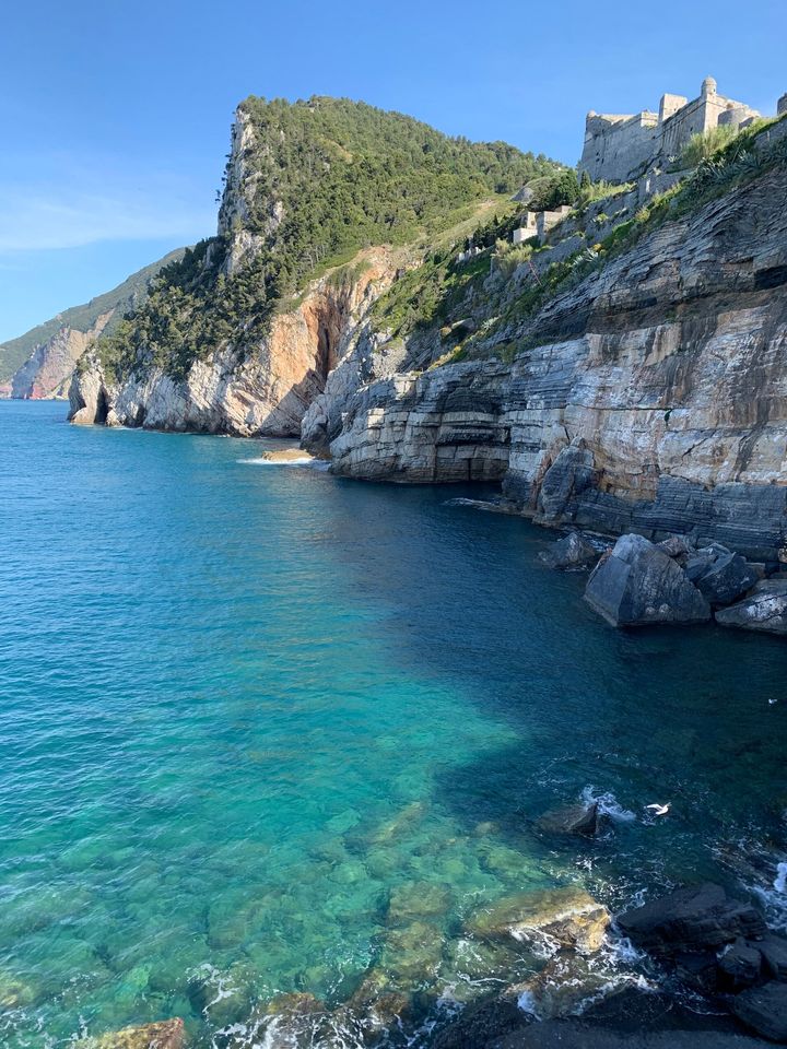 Mare azzurro contro una costa rocciosa, sormontata da fogliame verde e da una struttura in pietra sotto un cielo azzurro e terso.