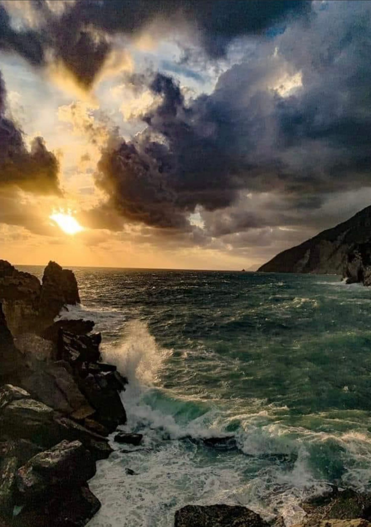 Le onde dell'oceano si infrangono contro la costa rocciosa sotto un cielo nuvoloso, mentre la luce del sole filtra.