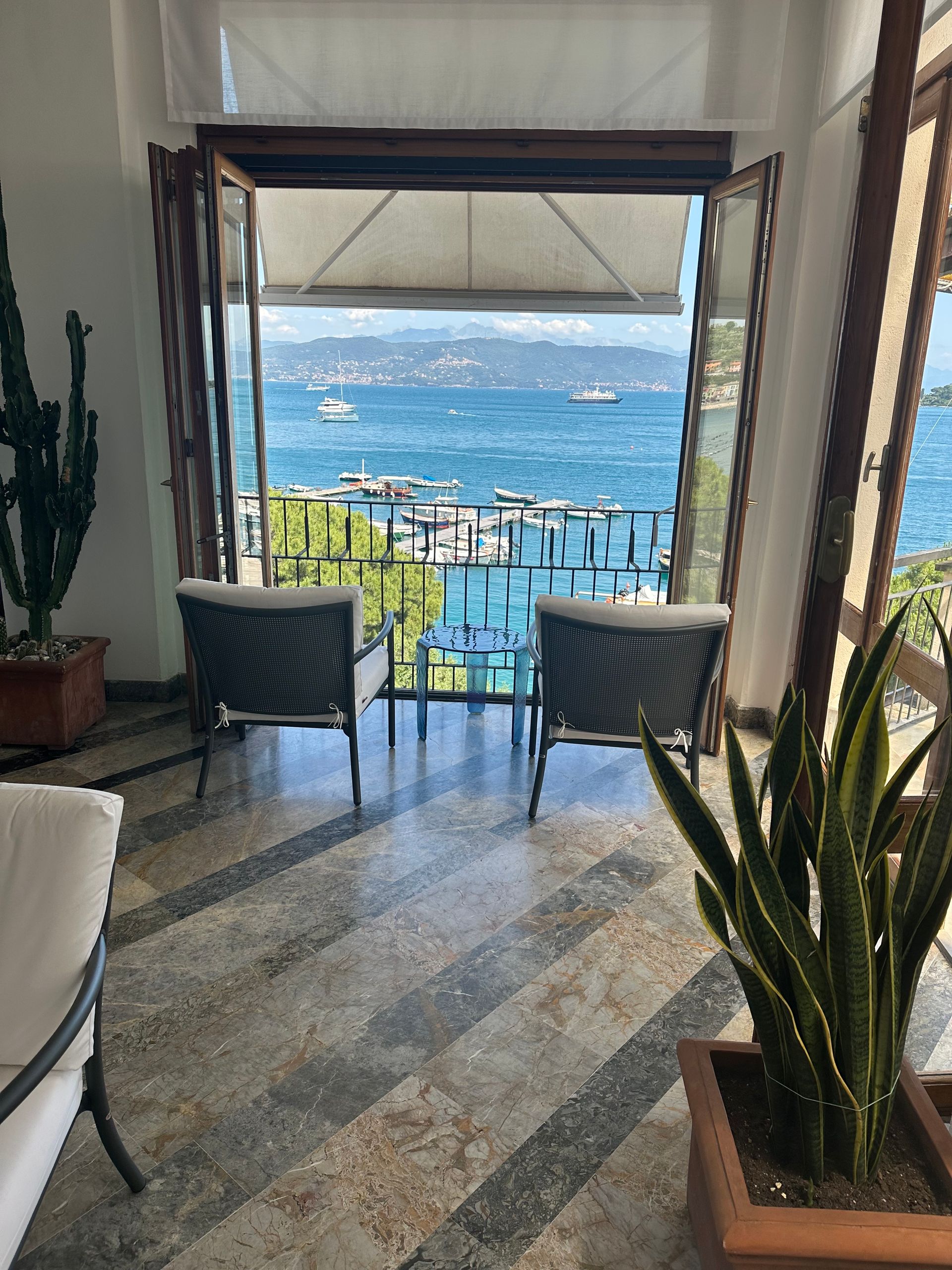 Le porte aperte rivelano una vista sul mare azzurro, con due sedie sul balcone. Le piante incorniciano l'interno con un pavimento piastrellato.