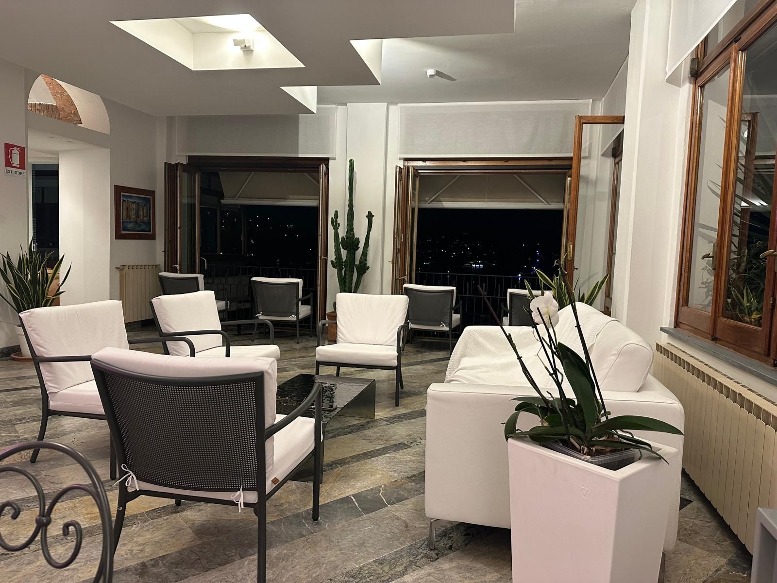 Area lounge con sedie bianche, tavolini e piante, con vista sull'esterno buio visibile attraverso le finestre.