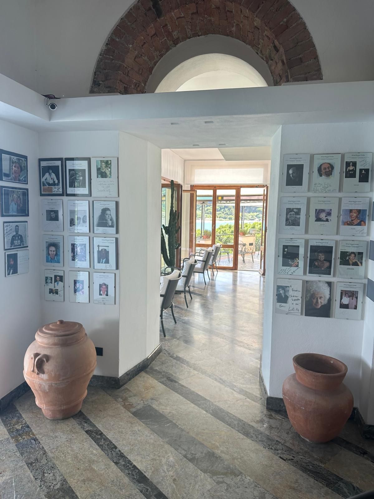 Corridoio con foto incorniciate su pareti bianche, vasi di terracotta, porta ad arco che conduce a una stanza piena di luce.