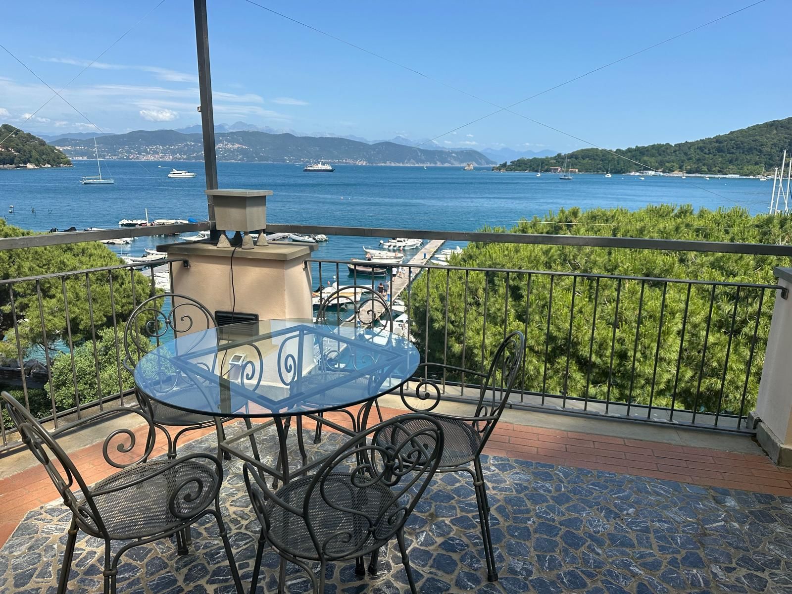 Balcone con vista sul mare blu, tavolo e sedie in vetro, porto e colline in lontananza.