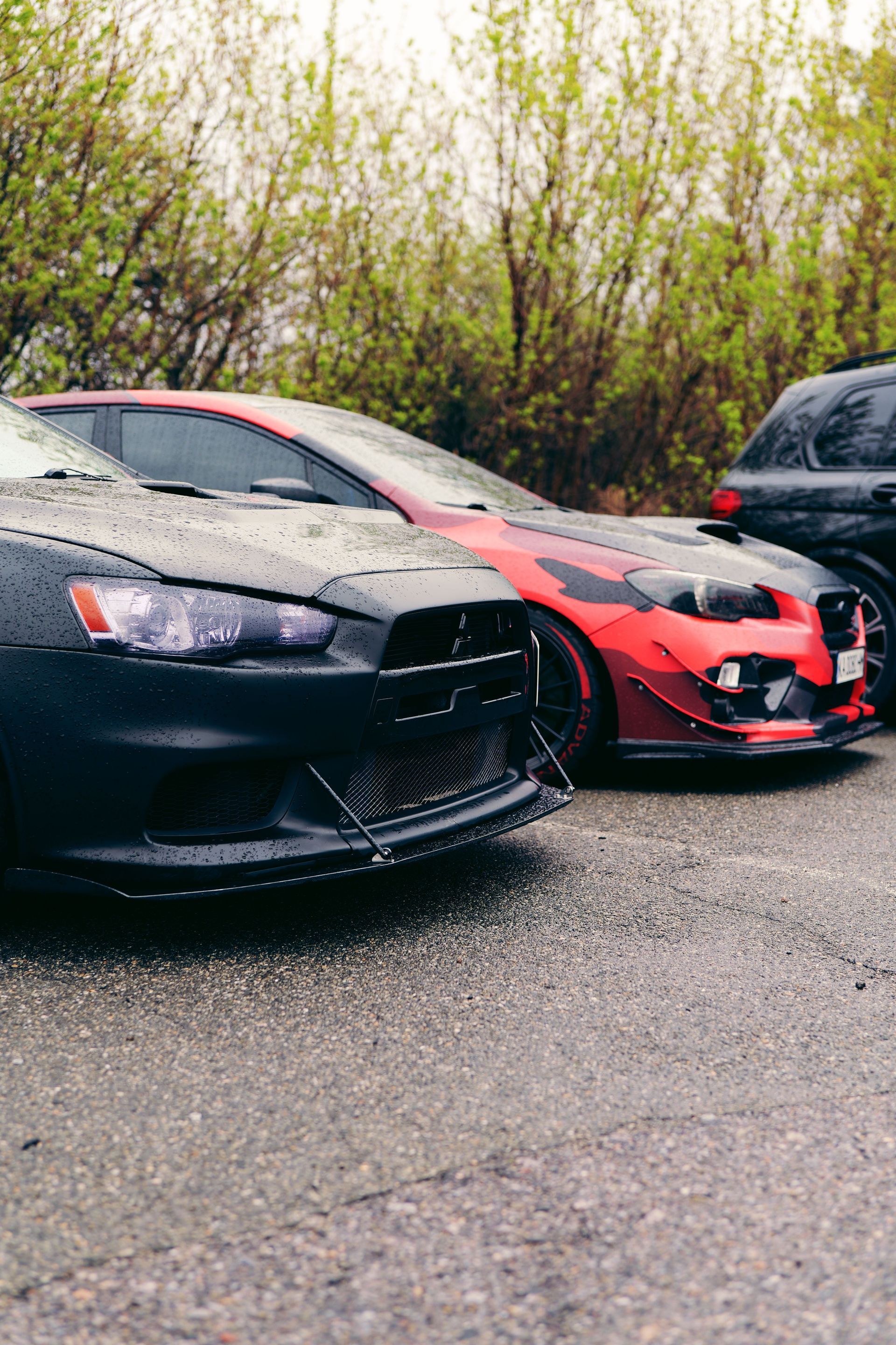 Two modified sports cars, black and red, parked on a wet roadside with trees behind them