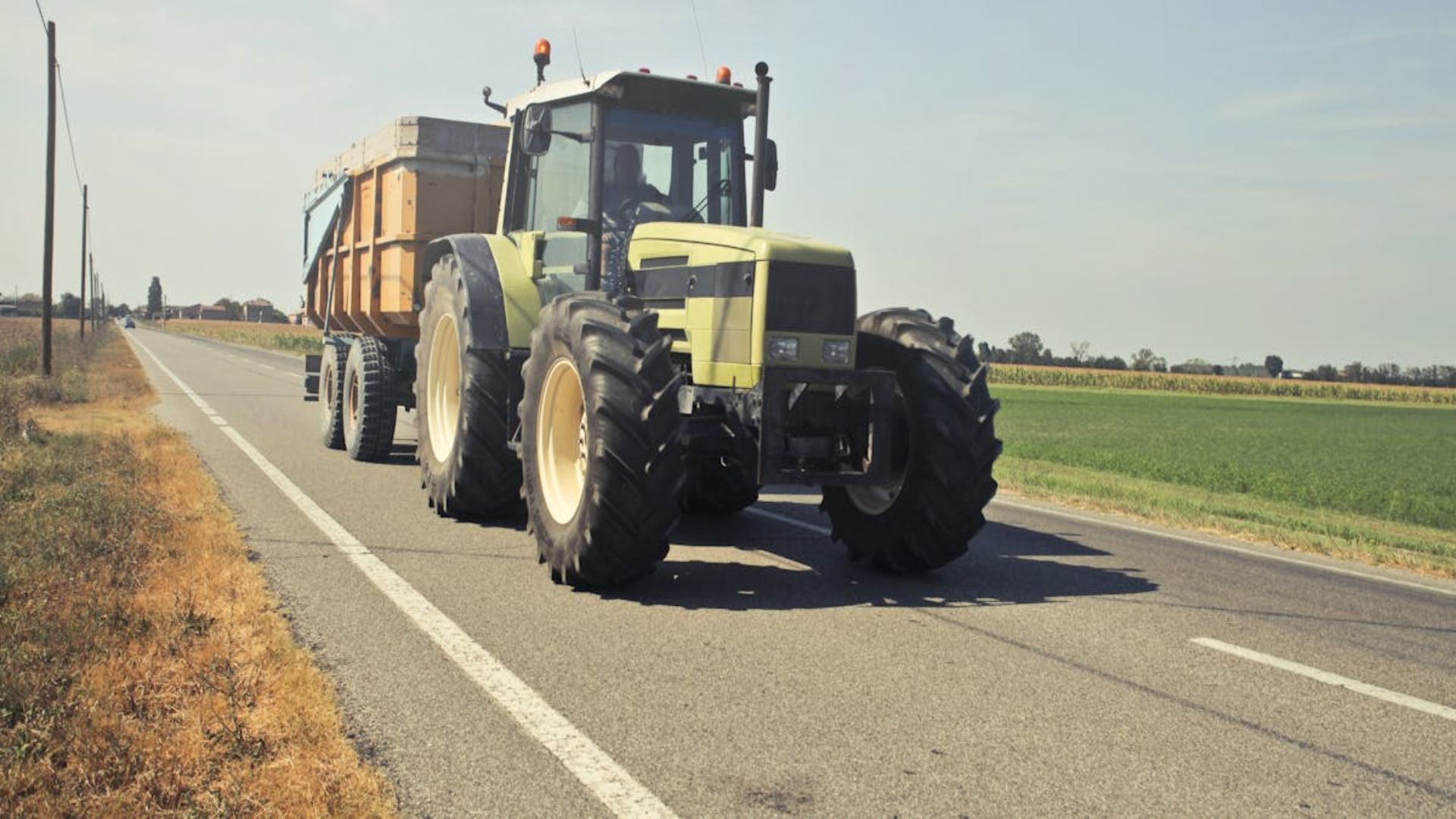 Tractor tirando de un remolque por una carretera asfaltada junto a un campo. Día soleado.