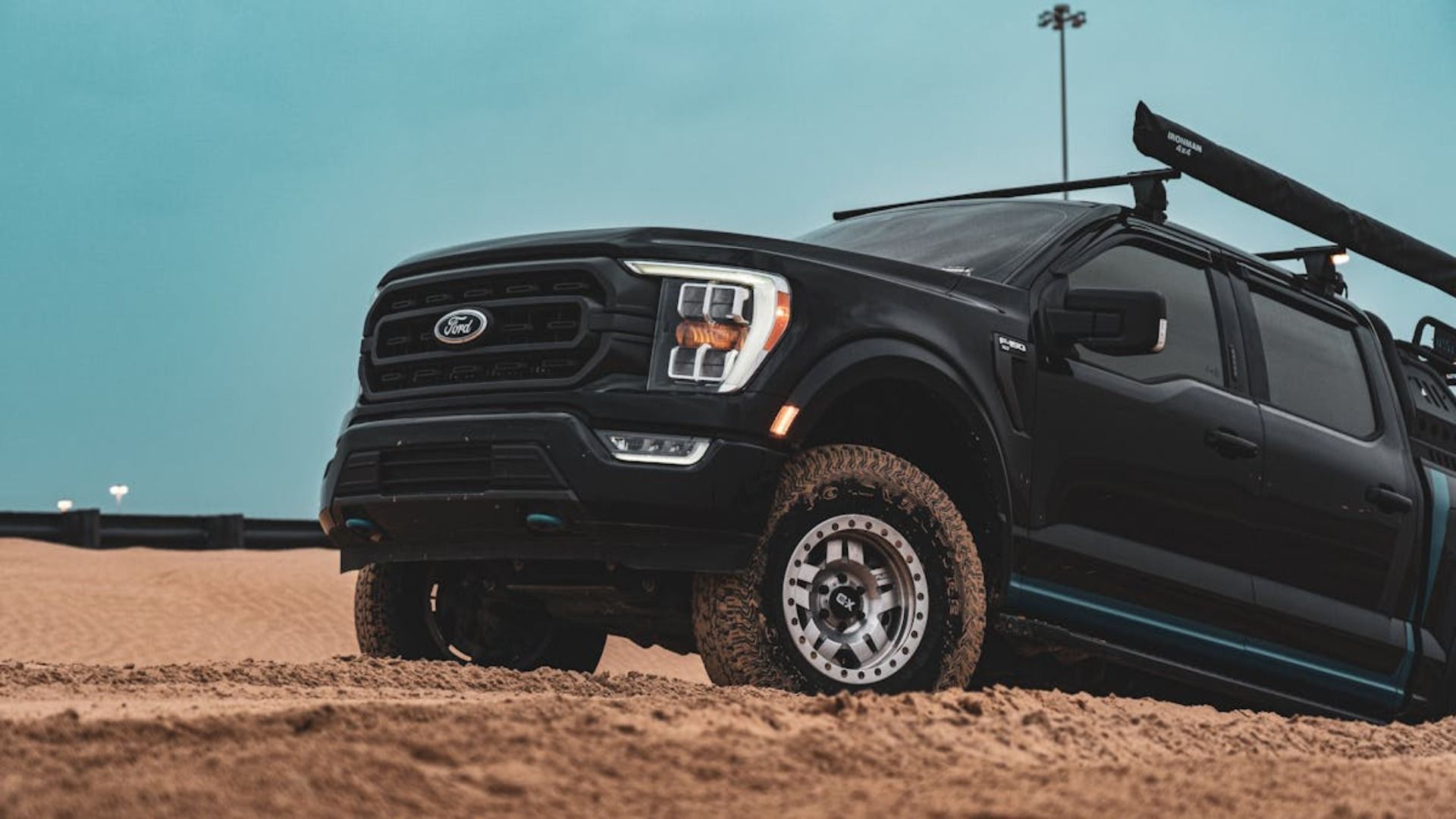 Camioneta Ford F-150 negra sobre arena, modificada con portaequipaje, toldo y neumáticos robustos. Cielo nublado.