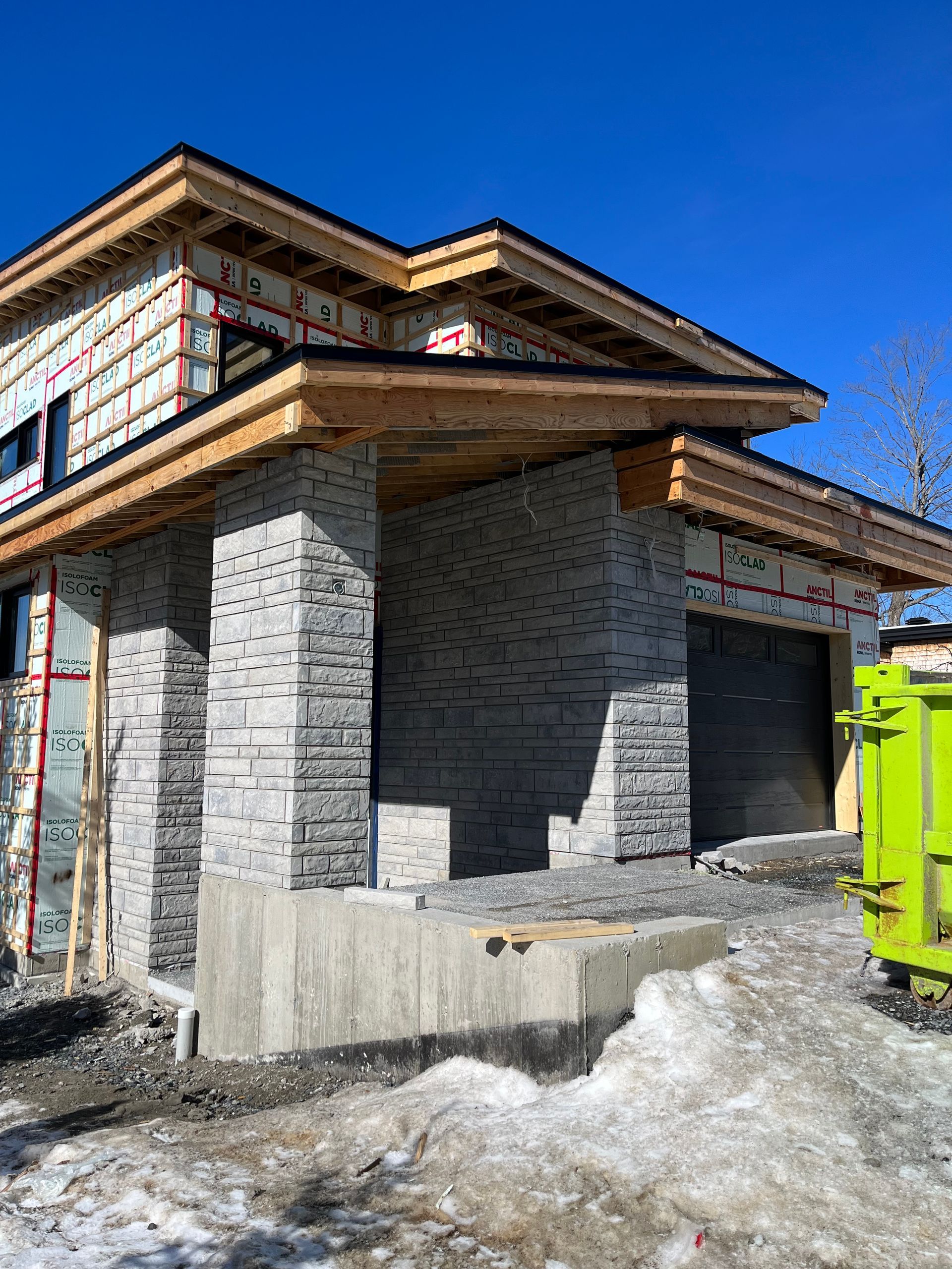 Une maison est en construction dans la neige avec une benne verte devant elle.
