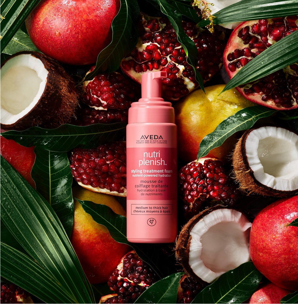 A pink Aveda hair product bottle surrounded by natural ingredients like coconuts &pomegranates. 