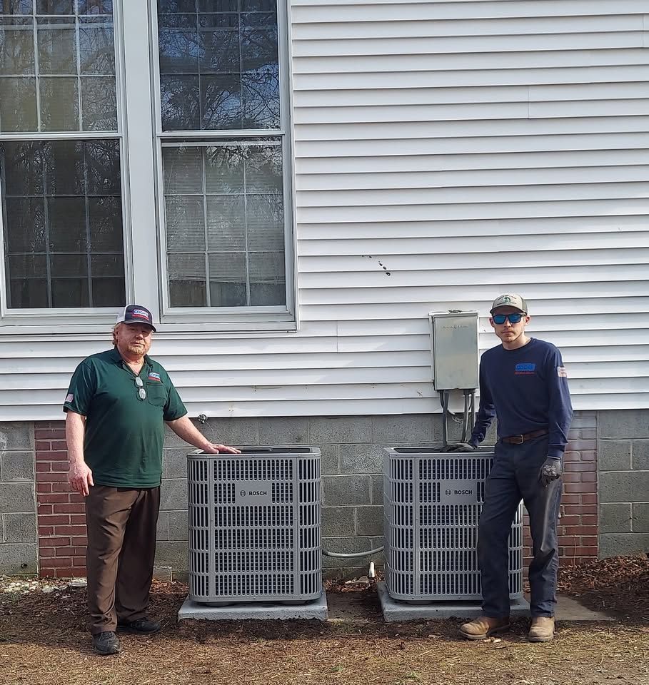 Two men are standing next to two air conditioners in front of a house.
