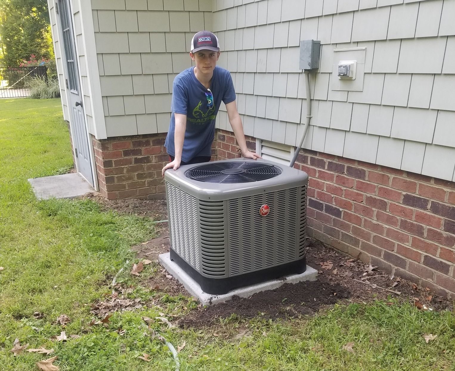A young man is standing next to an air conditioner outside of a house.