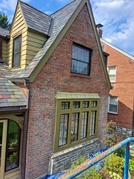 Brick house with a gabled roof and green-trimmed windows. A portion of a blue railing and plants are in the foreground.