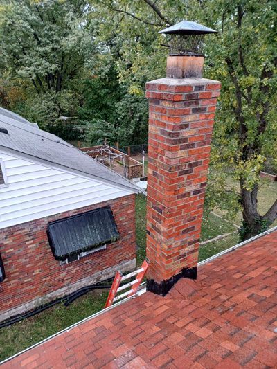 Brick chimney on a red shingled roof, with a metal cap.  A ladder leans against the chimney near a brick wall.