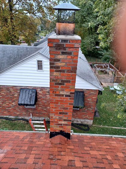 Brick chimney extending from a red-shingled roof, with a metal chimney cap and surrounded by a residential setting.