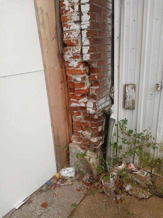 Damaged brick building corner; bricks are crumbling and painted white, with debris on the ground.