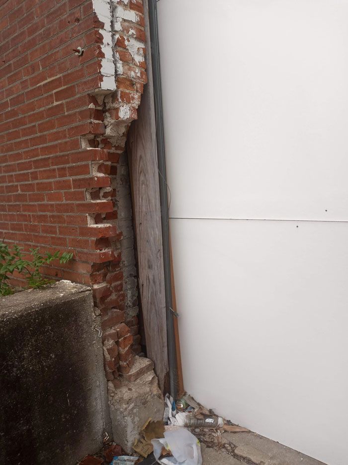 Damaged brick wall next to a white wall. Bricks are crumbling, revealing a wooden board. Concrete and debris are at the base.