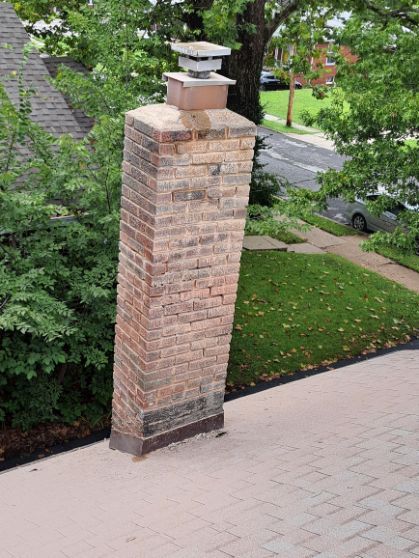 A brick chimney leaning to the side, on a roof, with trees and a street in the background. The chimney has a metal cap.