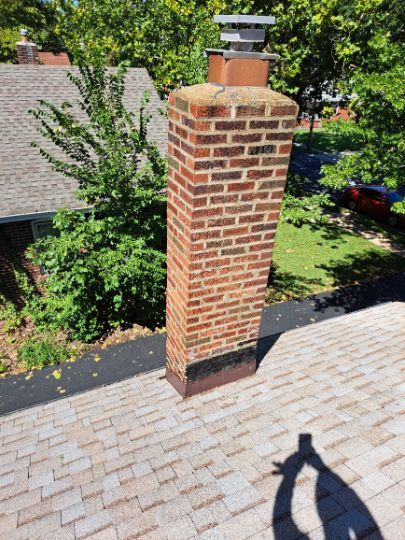 Brick chimney on a shingled roof, with a metal cap. The setting is a sunny day in a residential area.