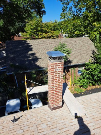Brick chimney with a metal cap on a brown shingled roof, scaffolding alongside. Green trees and blue sky in the background.