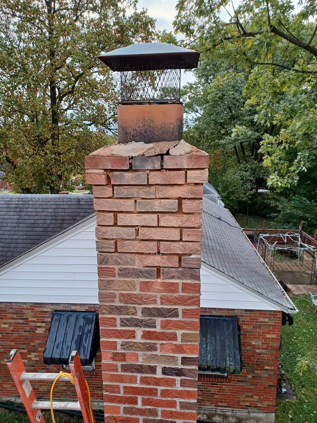 Brick chimney on a rooftop; deterioration visible at the top, topped with a chimney cap and surrounded by trees.