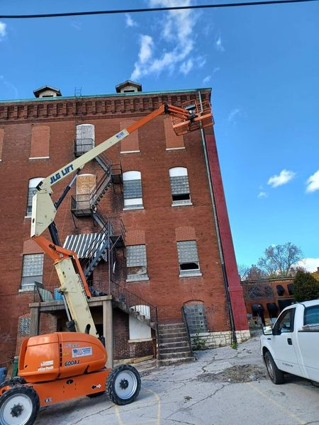 An orange boom lift extends toward a red brick building with boarded-up windows. A white truck is parked nearby.