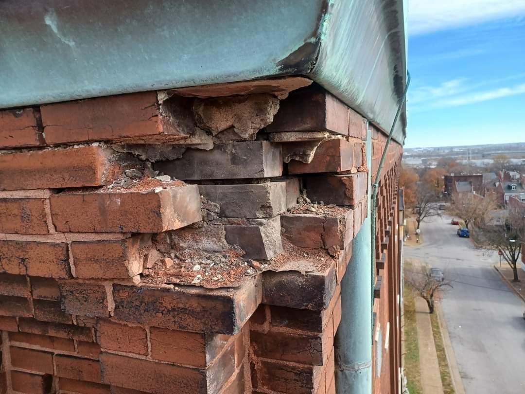 Damaged brick corner of a building, with crumbling bricks and exposed mortar, beneath a green-colored metal trim.