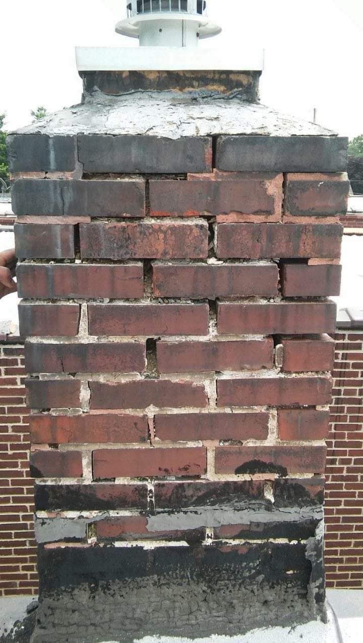 Brick chimney with significant mortar damage and black staining, atop a red brick building.