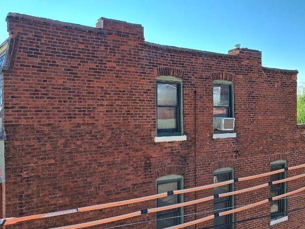 Brick building exterior with several windows, a small air conditioning unit, and a railing in the foreground; blue sky.