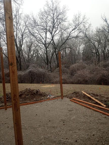 Construction site with wooden posts in the ground. Ground is gravel, surrounding area has dirt piles and scattered wood. Trees in the background.